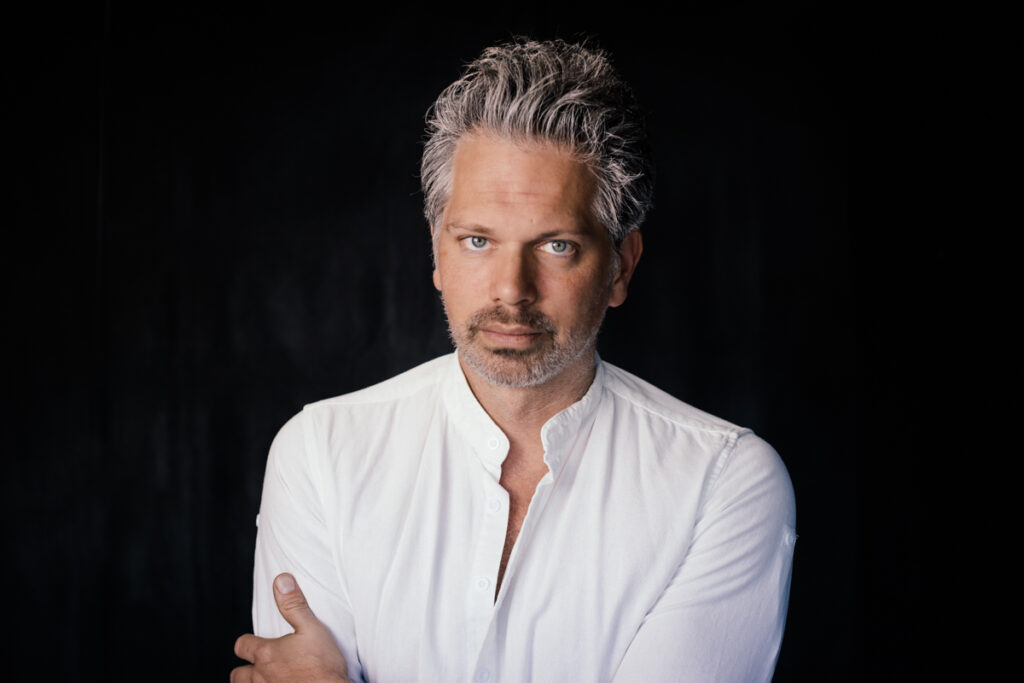 Portrait of Nick Holmes in a white shirt against a black backdrop, looking directly into camera